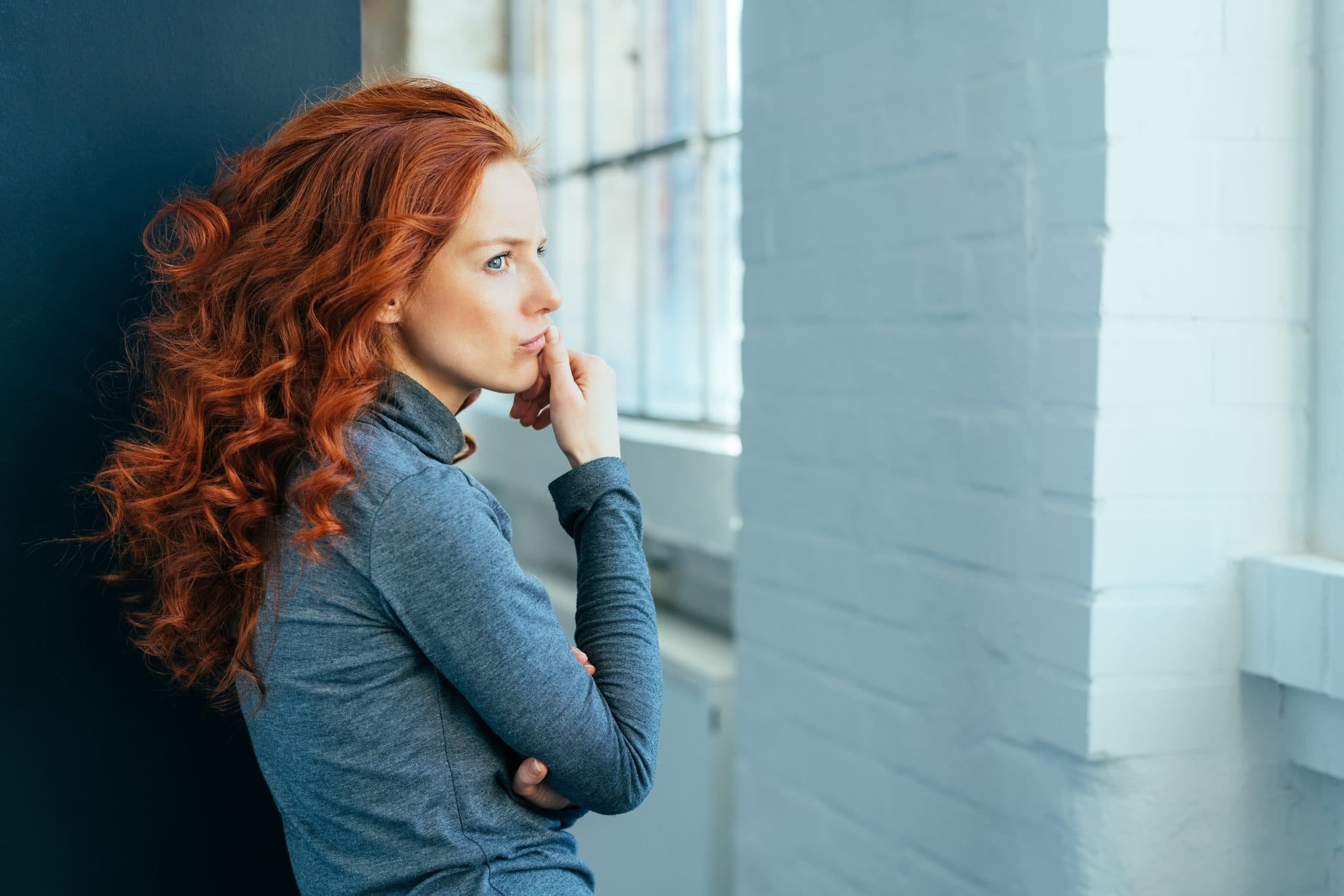 Sad lonely thoughtful young woman with gorgeous long curly red hair standing sideways indoors staring though a window