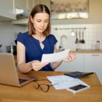 Check Your CSA Income Estimate. Accompanying image: thoughtful young woman using a laptop computer sitting at her kitchen holding utility bill and bank statements.
