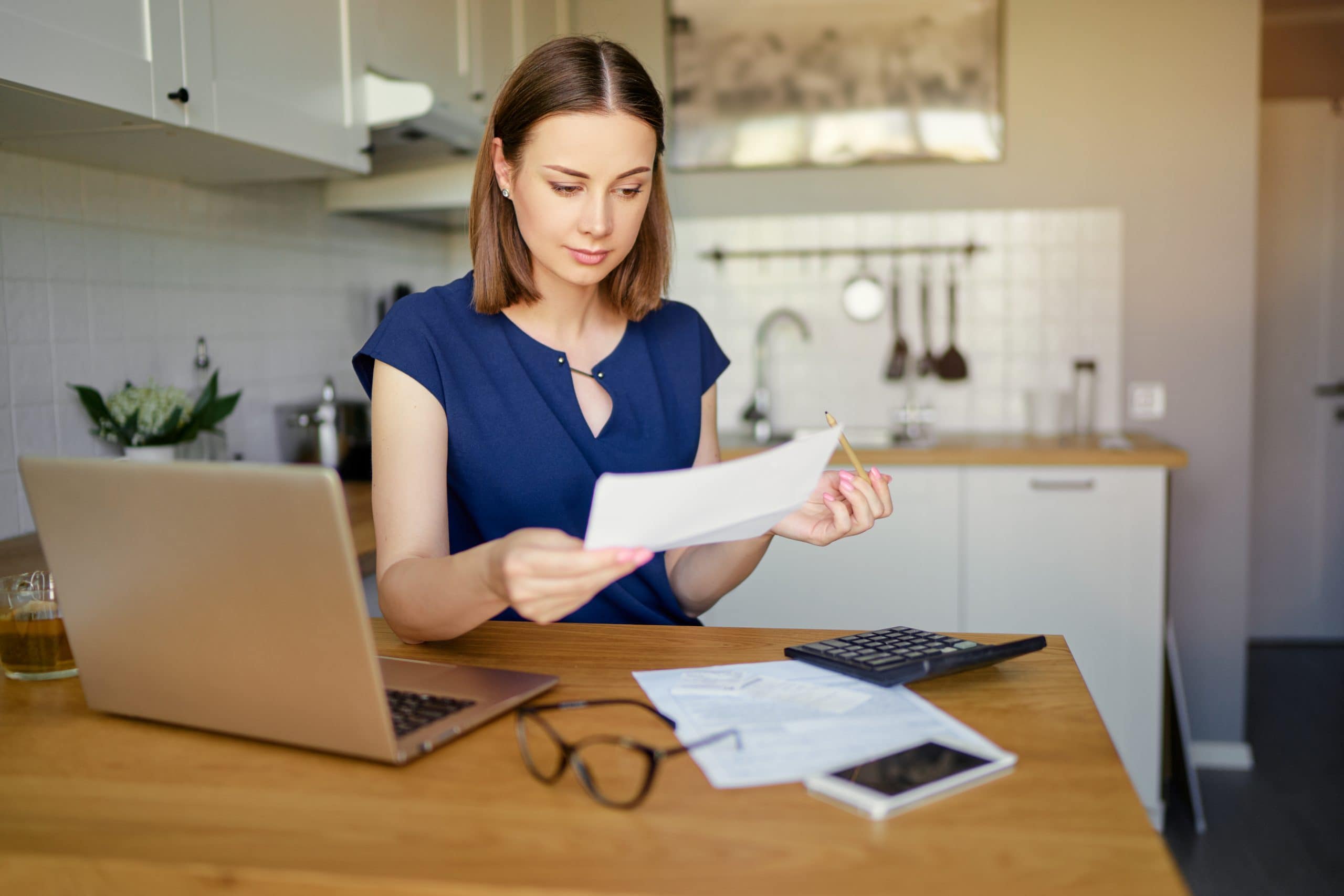Check Your CSA Income Estimate. Accompanying image: thoughtful young woman using a laptop computer sitting at her kitchen holding utility bill and bank statements.