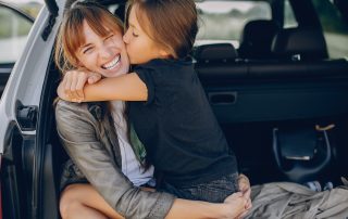 COVID-19 border closures – Can I still see my children? Accompanying picture: Fashionable mother with daughter. Family is sitting in the car trunk. Girl in a black t-shirt is hugging the mother.